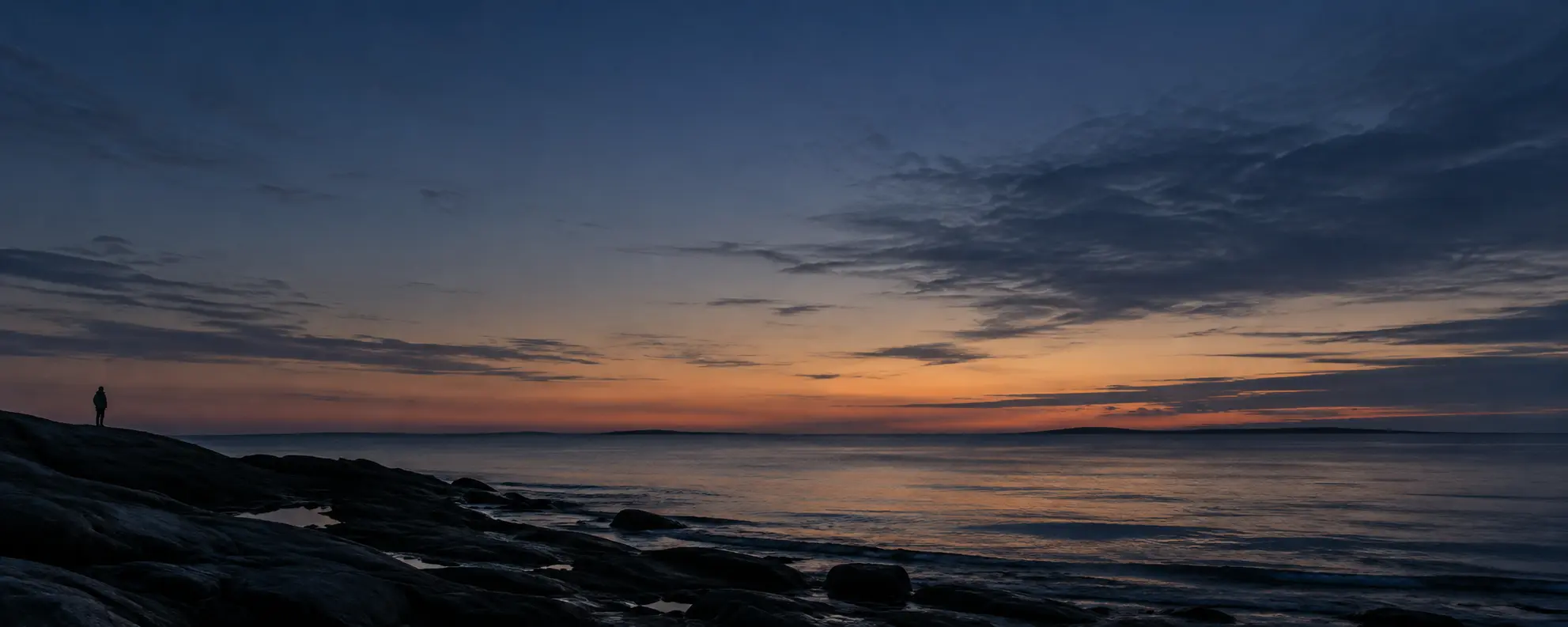 A lone figure stands by the water beneath the fading light of dusk